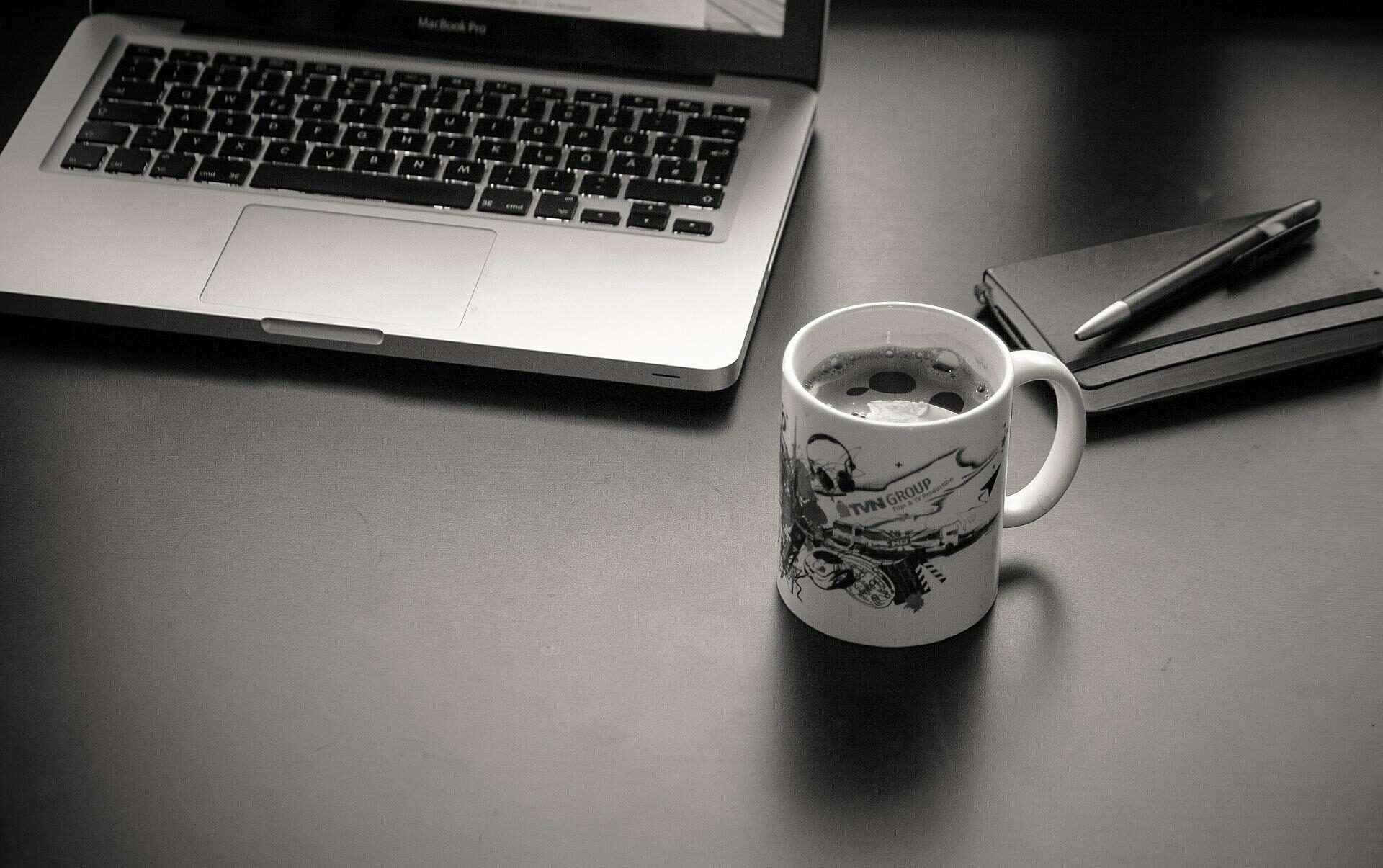 Laptop, Block und Stift und eine Kaffeetasse stehen auf einem Schreibtisch. Das Bild ist in schwarz weiß